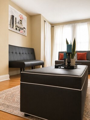 Ein Wohnzimmer mit schwarzem Sofa, schwarzem Hocker, einem Couchtisch mit Dekoration und einem Wandbild.