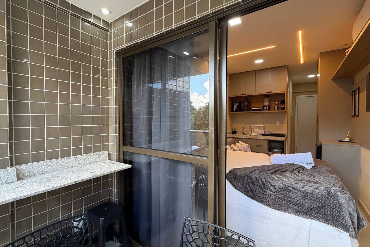 Una habitación de hotel moderna con un ventanal grande, una cama, una pequeña kitchenette y un rincón de descanso con una silla y una mesita.
