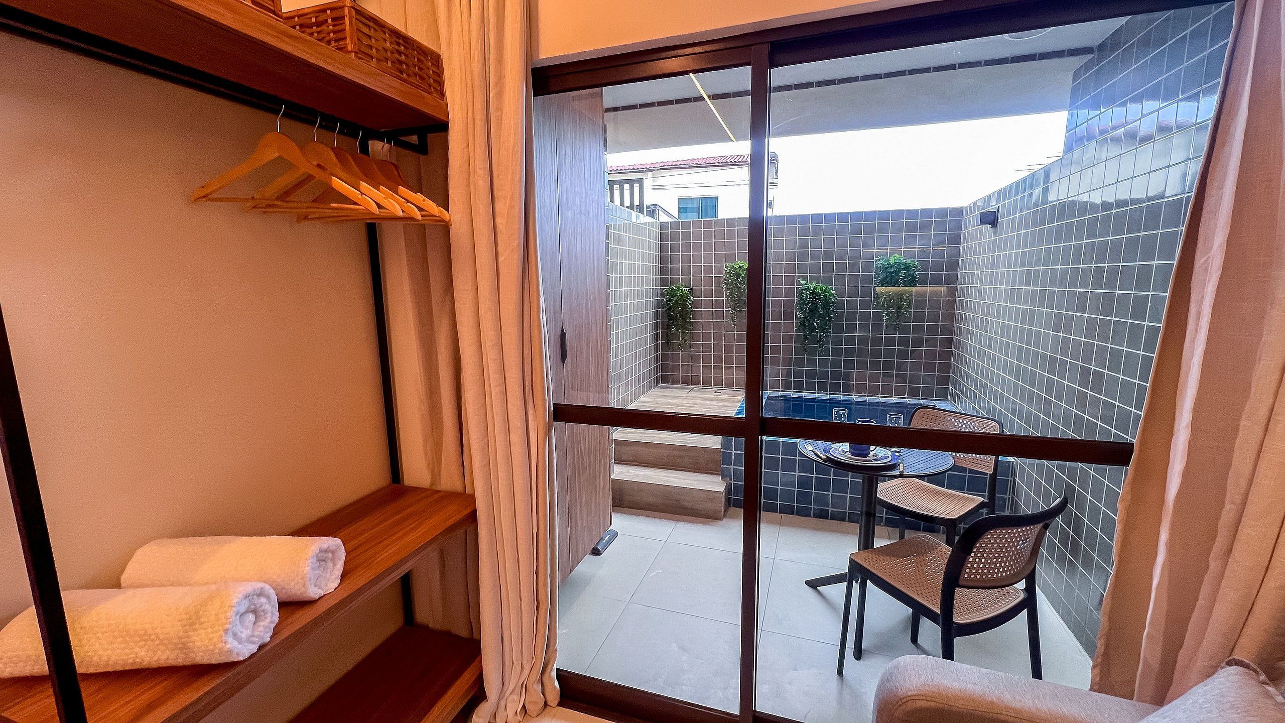 Un balcon avec une table et des chaises, une étagère en bois avec des serviettes et une vue sur une cour dallée.