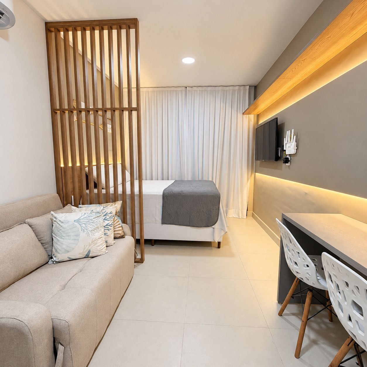 Habitación de hotel moderna con sofá, cama con manta gris, escritorio con sillas, televisor montado en la pared y un separador de madera.