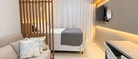 Habitación de hotel moderna con sofá, cama con manta gris, escritorio con sillas, televisor montado en la pared y un separador de madera.