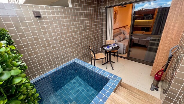 Una piscina pequeña al aire libre con borde de azulejos, una mesa con sillas y un ventanal corredero que da a un espacio interior.