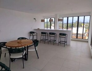 A spacious room with a round wooden table and six chairs, a small bar area with stools, and large windows offering a view of the outdoors.