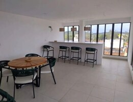 Una habitación amplia con una mesa redonda de madera y seis sillas, una pequeña zona de bar con taburetes y ventanas grandes que ofrecen vistas al exterior.