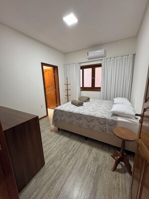 A bedroom with a bed, a wooden desk, a window with curtains, and an air conditioning unit. - COZY HOUSE COMPLETE IN ARRAIAL DAJUDA (PORTO SEGURO)