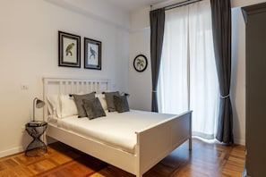 A bedroom with a white bed, a bedside table with a lamp, and a window with curtains.