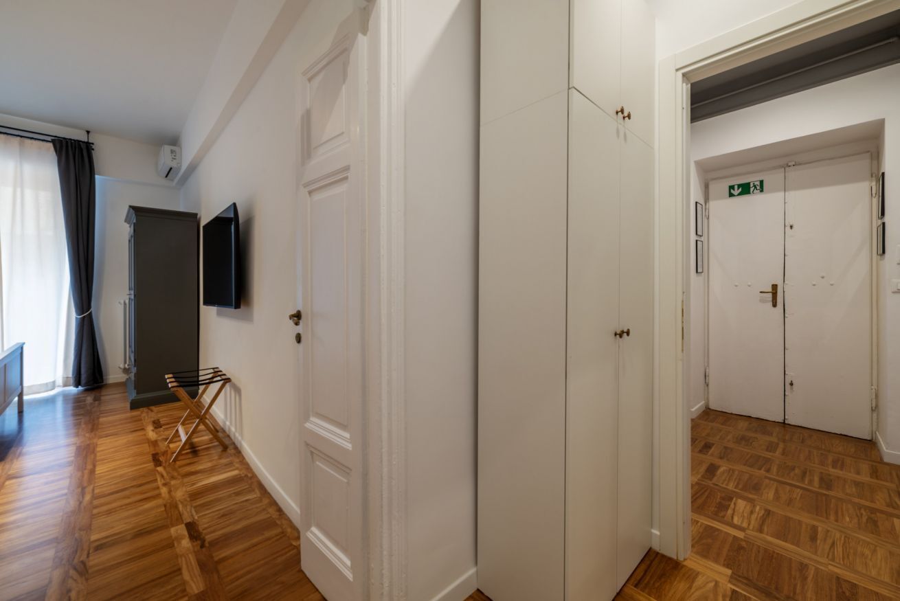 A hallway with wooden flooring, white doors, a flat-screen TV mounted on the wall, and a staircase leading to a door marked with an emergency exit sign.