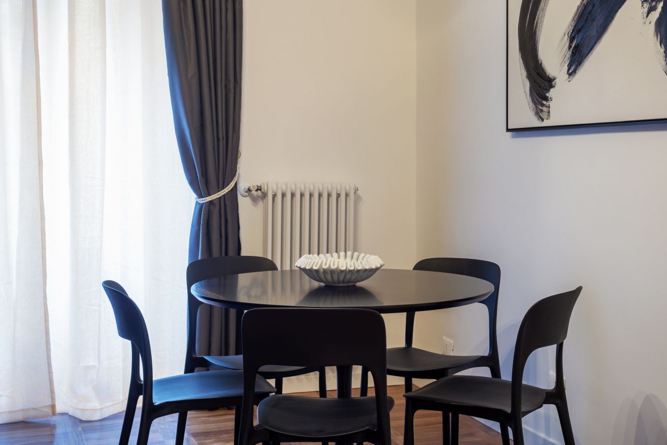 Zona pranzo con tavolo rotondo, quattro sedie, una ciotola bianca, un termosifone e un quadro appeso al muro.