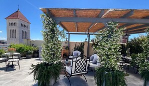 Un patio al aire libre con mobiliario, una pérgola y una torre al fondo.