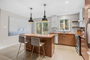 Una cocina moderna con una isla central, taburetes de bar, iluminación colgante y un ventanal grande.