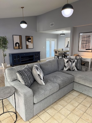 Un salon moderne avec un canapé d’angle gris, des coussins aux motifs noir et blanc, un meuble de rangement pour appareils électroniques de couleur bleu foncé, et un coin repas à l’arrière-plan.