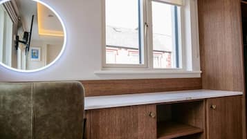 Une pièce avec un meuble en bois, une fenêtre avec des stores, un miroir rond avec un éclairage et une chaise.