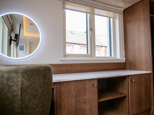 Ein Zimmer mit Holzschrank, Jalousiewindfang und rundem Spiegel mit Beleuchtung, sowie einem Sessel.