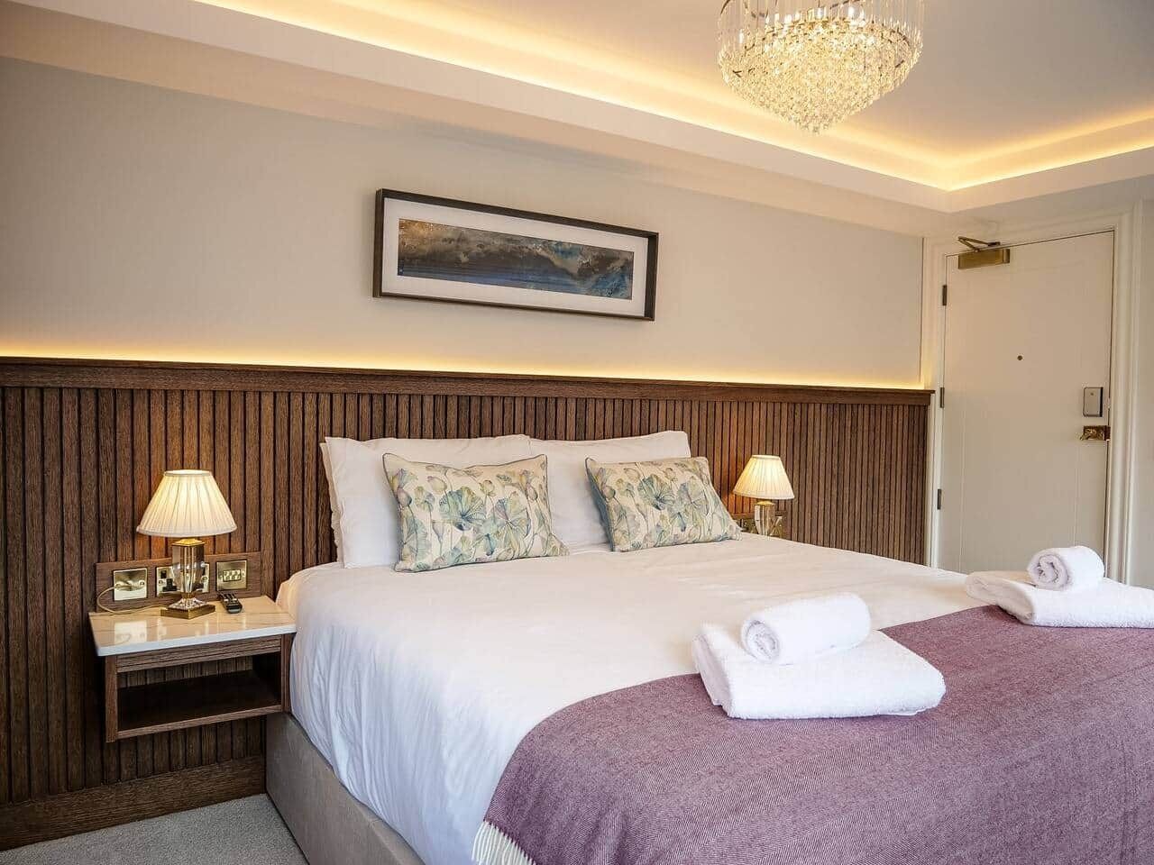 Une chambre d’hôtel avec une tête de lit en bois, un grand lit avec une literie blanche, deux lampes de chevet, un tableau encadré au mur et un lustre.