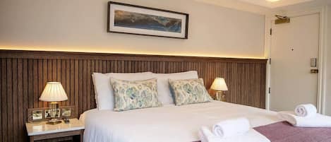 Une chambre d’hôtel avec une tête de lit en bois, un grand lit avec une literie blanche, deux lampes de chevet, un tableau encadré au mur et un lustre.