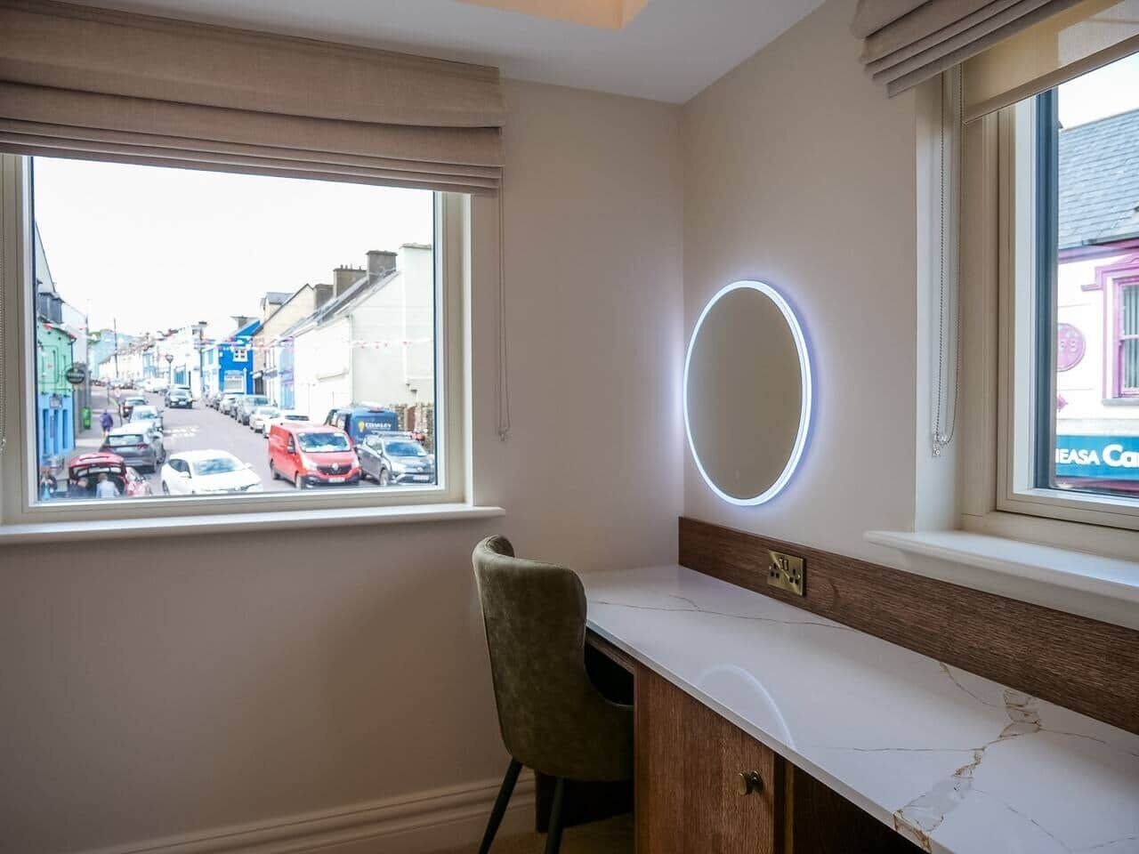 A room with a vanity, a chair, a window with a view of a street, and a circular mirror.