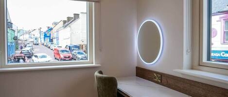 A room with a vanity, a chair, a window with a view of a street, and a circular mirror.
