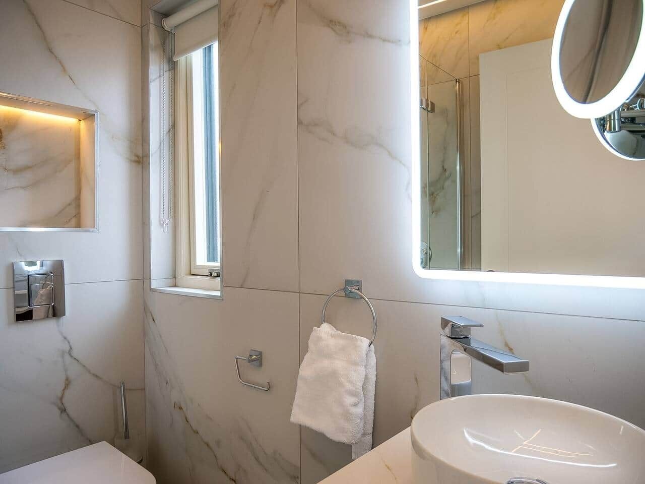 Un baño con pared de mármol, una ventana, toallero, lavabo y espejo.