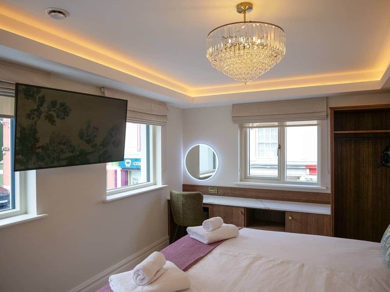 A hotel room with a bed, a desk, a chair, a round mirror, and a chandelier.