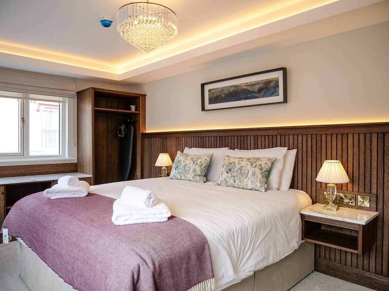 Habitación de hotel con una cama grande, dos mesitas de noche, un candelabro, una ventana y un cuadro en la pared.