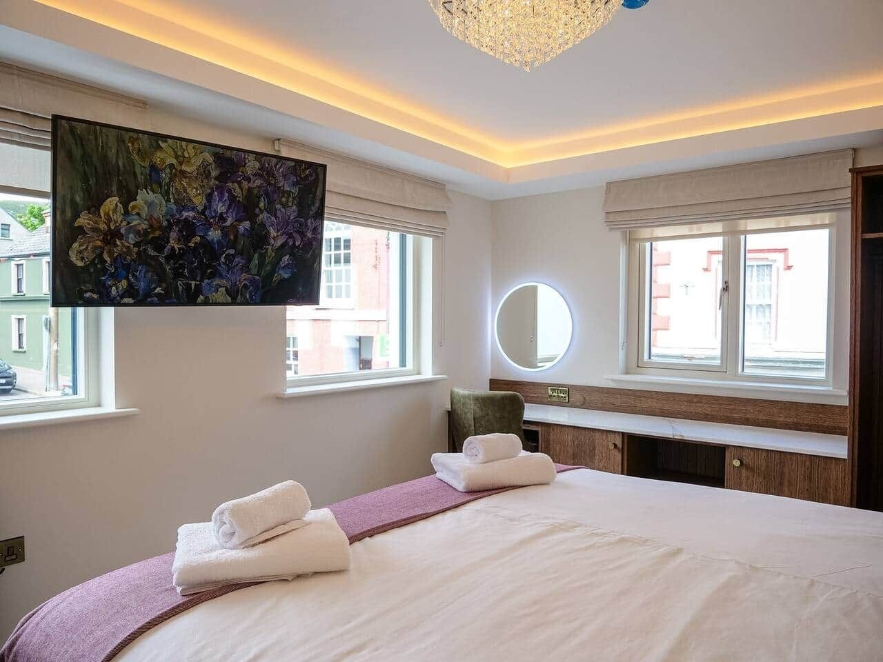 A hotel room with a large bed, a round mirror, a painting, and a view of buildings outside.