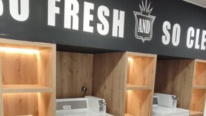 Ein Waschraum mit mehreren Waschmaschinen und Trocknern, Holzregalen und einem Schild mit der Aufschrift 'SO FRESH AND SO CLEAN'.