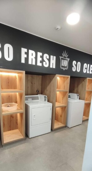 Ein Waschraum mit mehreren Waschmaschinen und Trocknern, Holzregalen und einem Schild mit der Aufschrift 'SO FRESH AND SO CLEAN'.