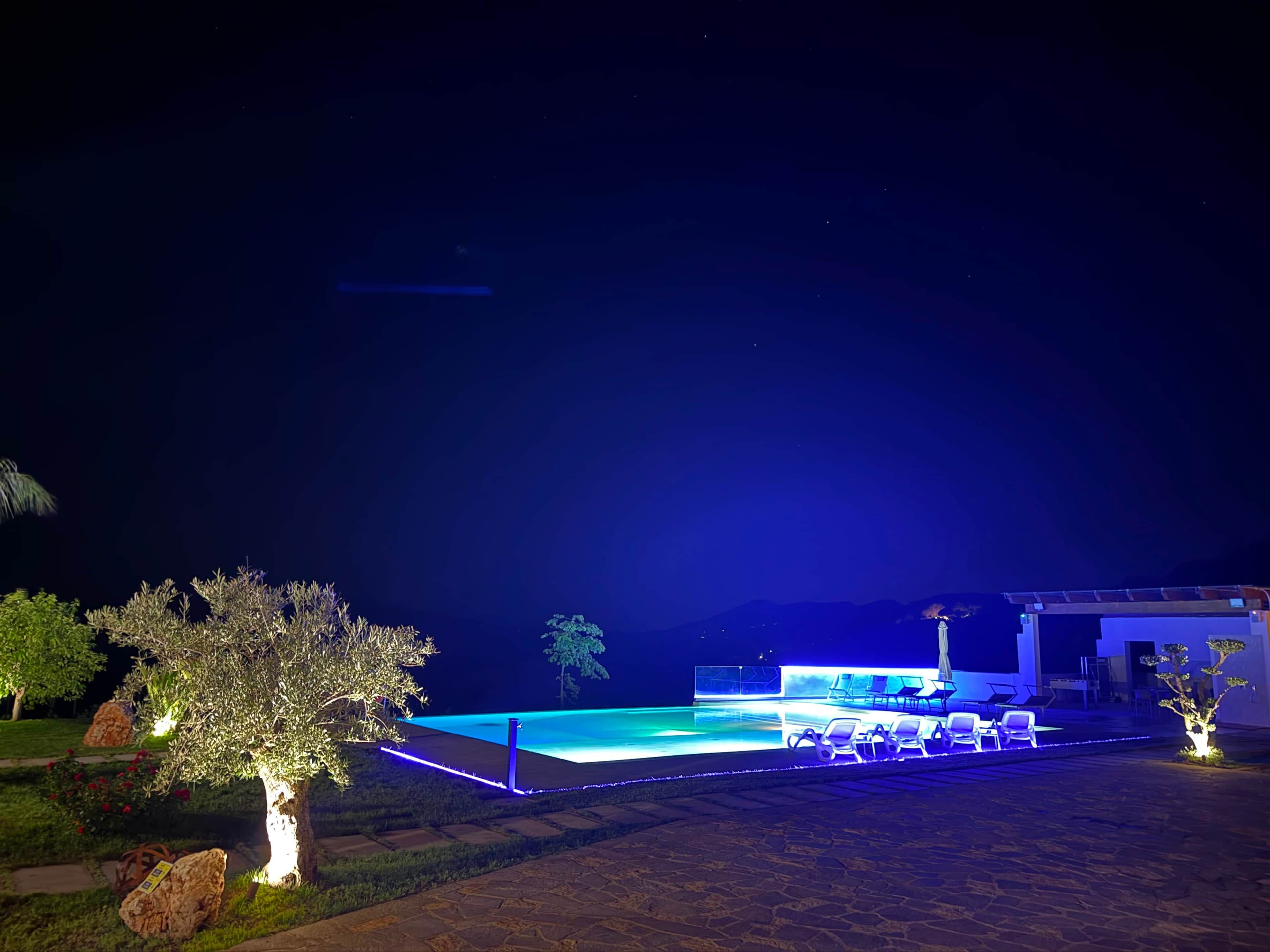 Un espace extérieur avec piscine, aménagé pour la nuit, doté d’une surface plane, de chaises longues et d’un bâtiment en arrière-plan.