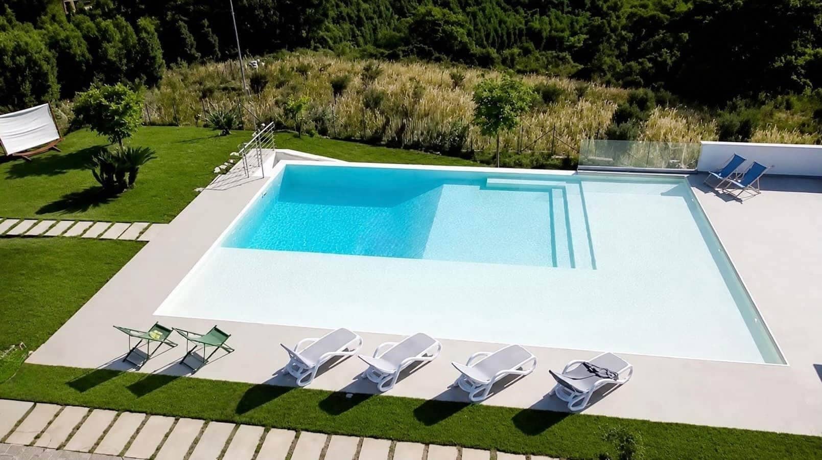 Una piscina rettangolare con acqua cristallina circondata da una terrazza pavimentata. Diverse sedie a sdraio bianche sono disposte lungo il bordo della piscina.