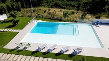 Una piscina rettangolare con acqua cristallina circondata da una terrazza pavimentata. Diverse sedie a sdraio bianche sono disposte lungo il bordo della piscina.