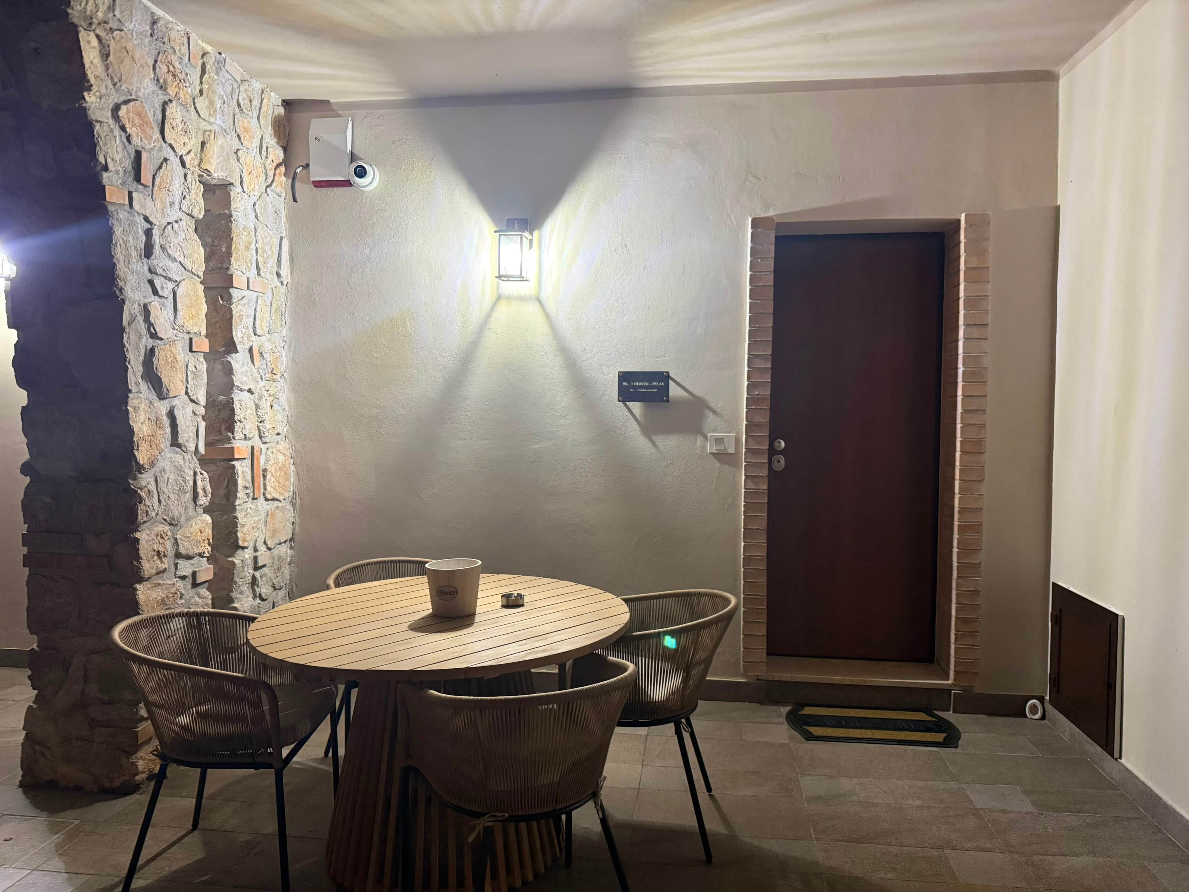 Ein Zimmer mit einer Steinsäule, einem runden Holztisch mit Stühlen, einer Tür und einer Wandleuchte.