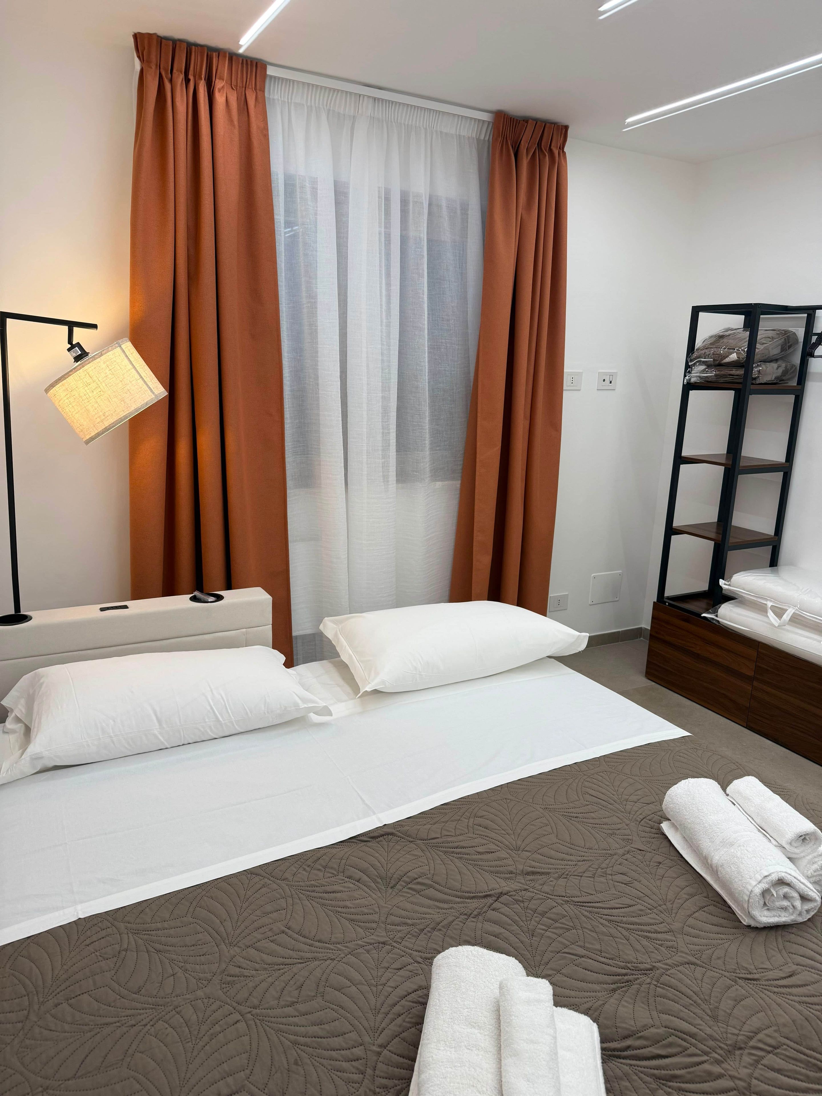 Une chambre d’hôtel avec un lit, une lampe de chevet, une étagère avec des serviettes et une fenêtre avec des rideaux.