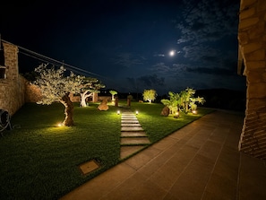 A well-lit outdoor pathway leading to a stone building, surrounded by landscaped greenery and illuminated trees.
