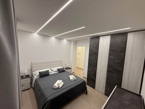 A modern hotel room with a bed, bedside tables, a nightstand, and a door leading to another room.