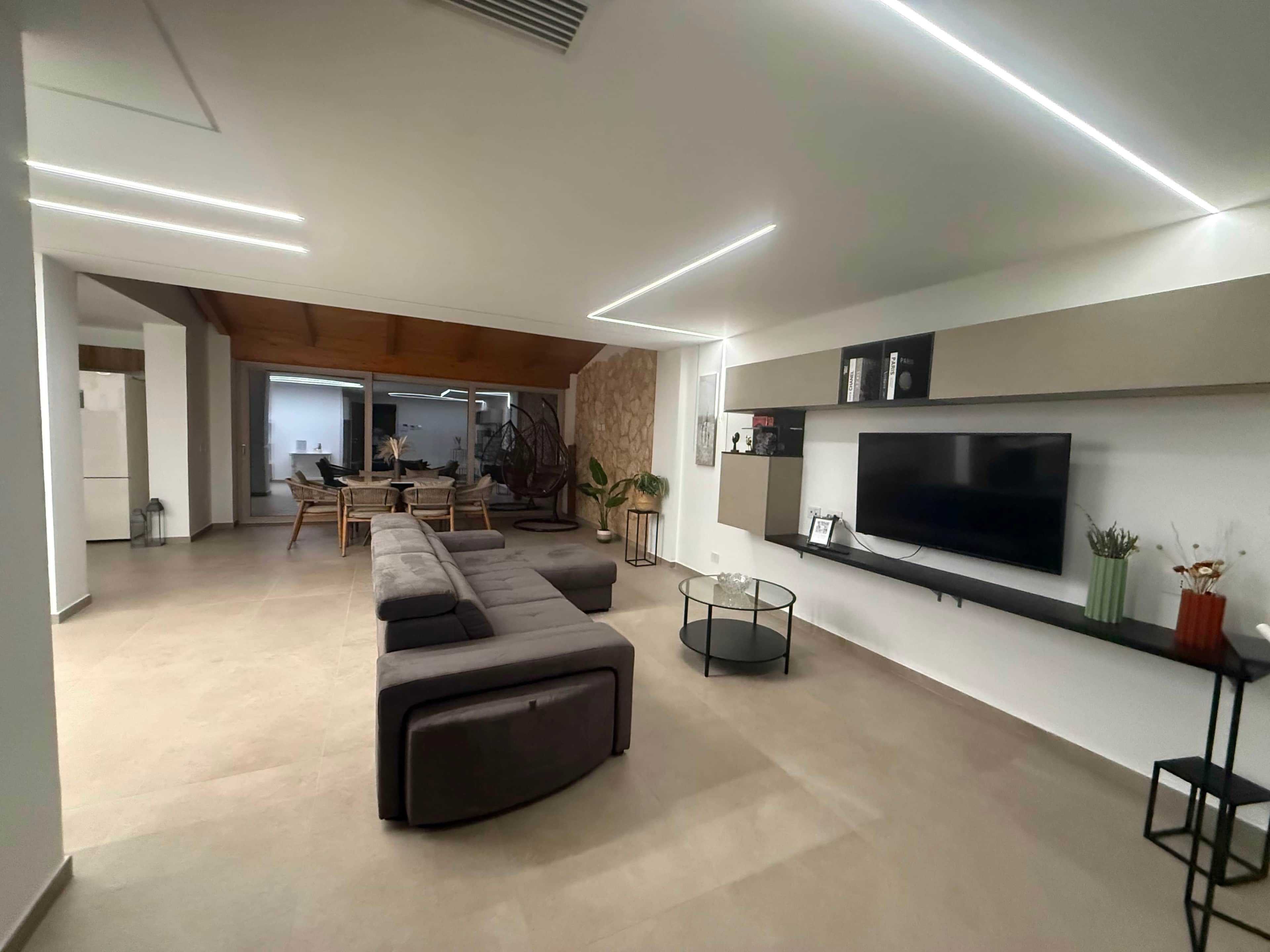 Een moderne woonkamer met een grote flatscreen televisie, een hoekbank en een eethoek met rieten stoelen.