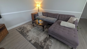 A living room with a sectional sofa, a coffee table with a plant, a lamp, and a wooden cabinet. - Entire House & Windmill Coast Retreat (Glace Bay)