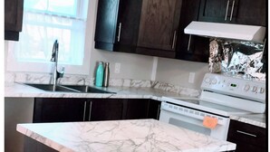 A kitchen with dark cabinets, a marble countertop island, a stove, and a window with natural light. - Entire House & Windmill Coast Retreat (Glace Bay)