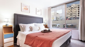 Een moderne hotelkamer met een groot bed, nachtkastjes, een raam met uitzicht op gebouwen en een balkon.