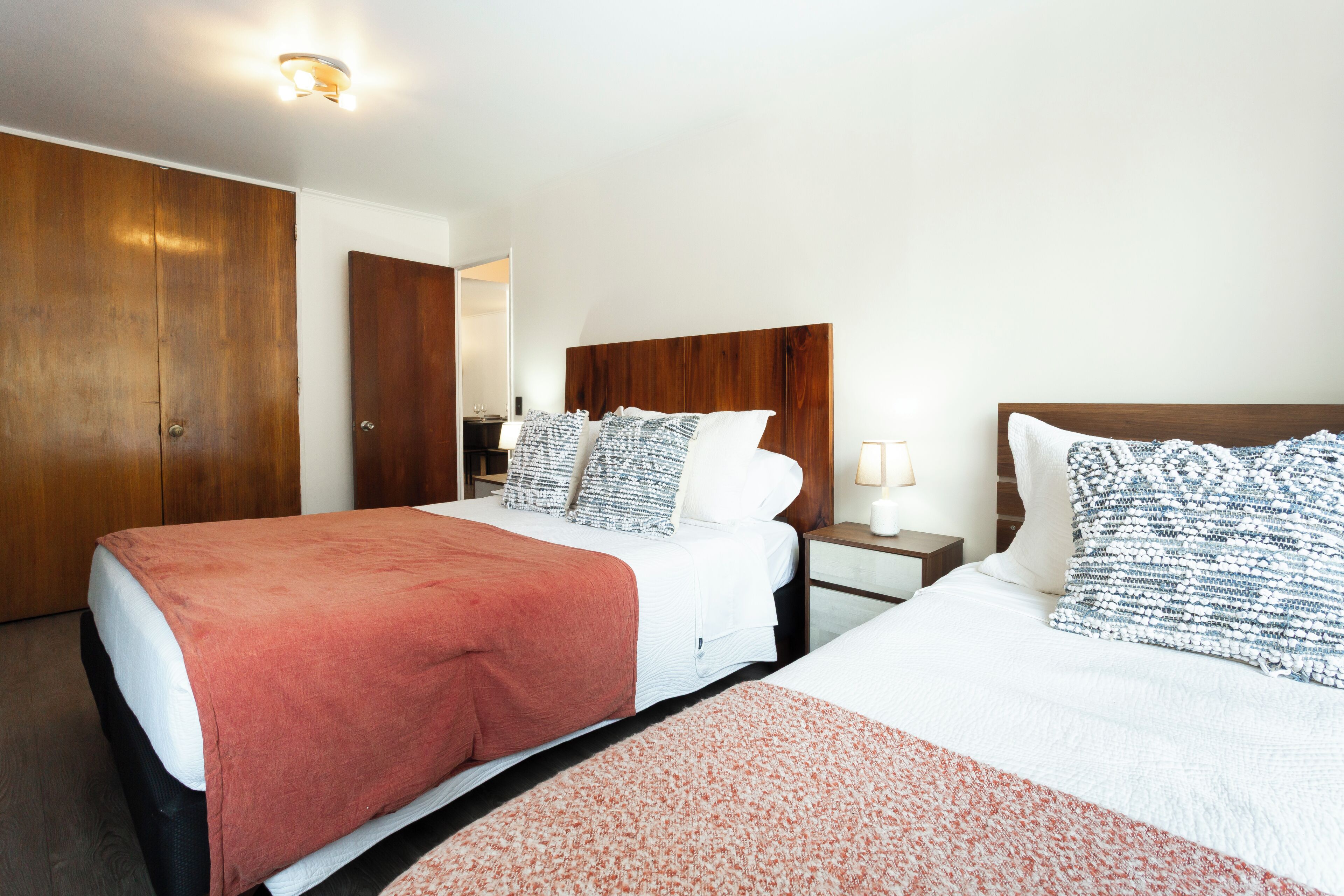 A hotel room with two beds, wooden headboards, a nightstand with a lamp, and a wardrobe.