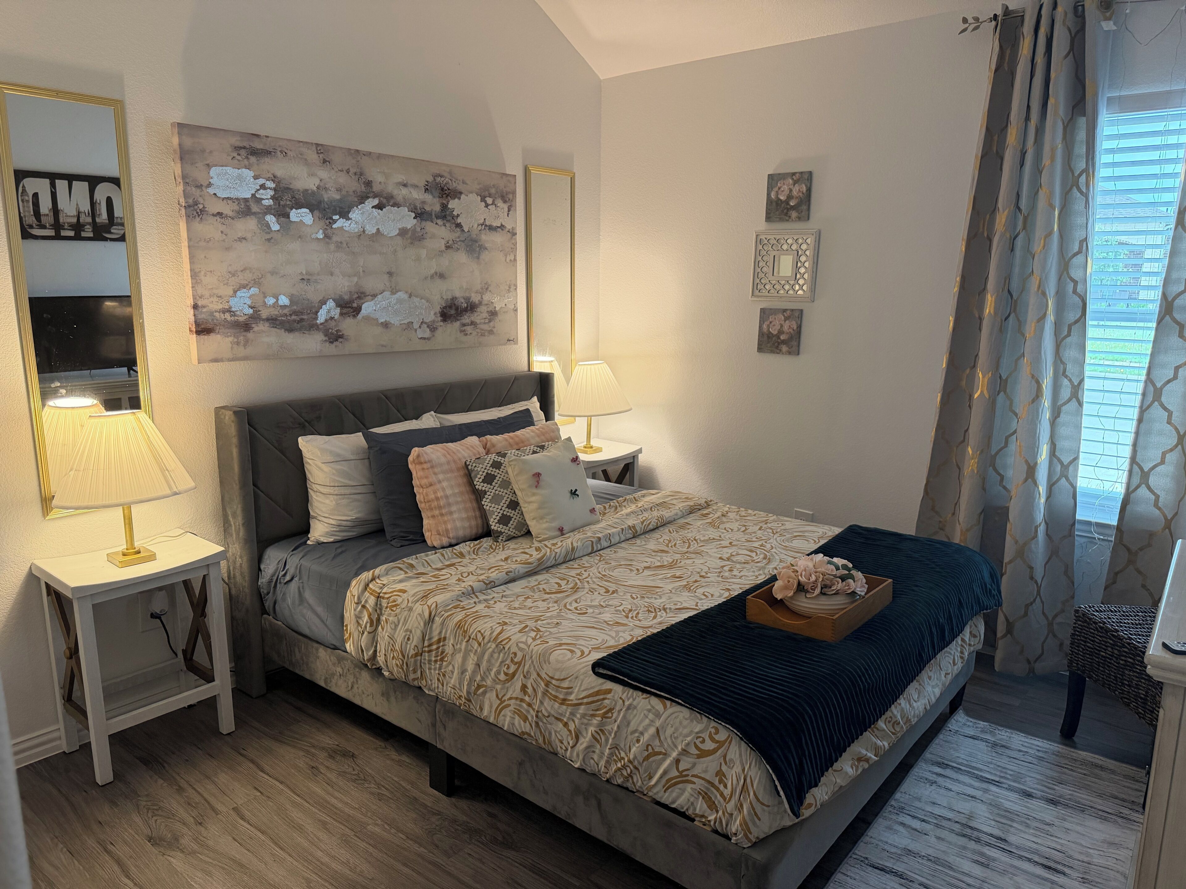 Un dormitorio con cama, mesitas de noche, lámpara, espejo y ventana con cortinas.