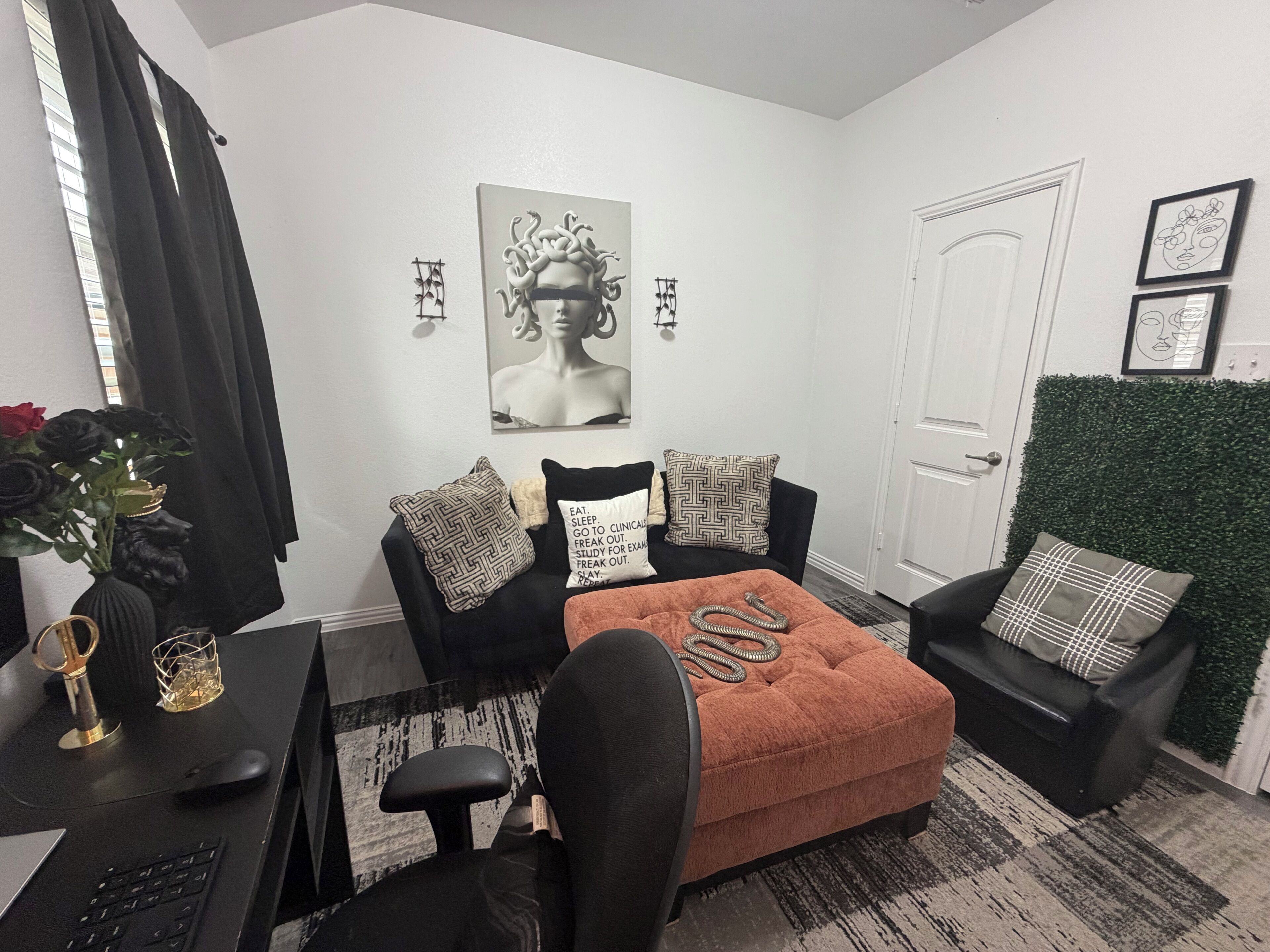 Een moderne woonkamer met een zwarte bank, een klein oranje poefje, een zwarte stoel, een bureau met een computer en een vaas met bloemen.
