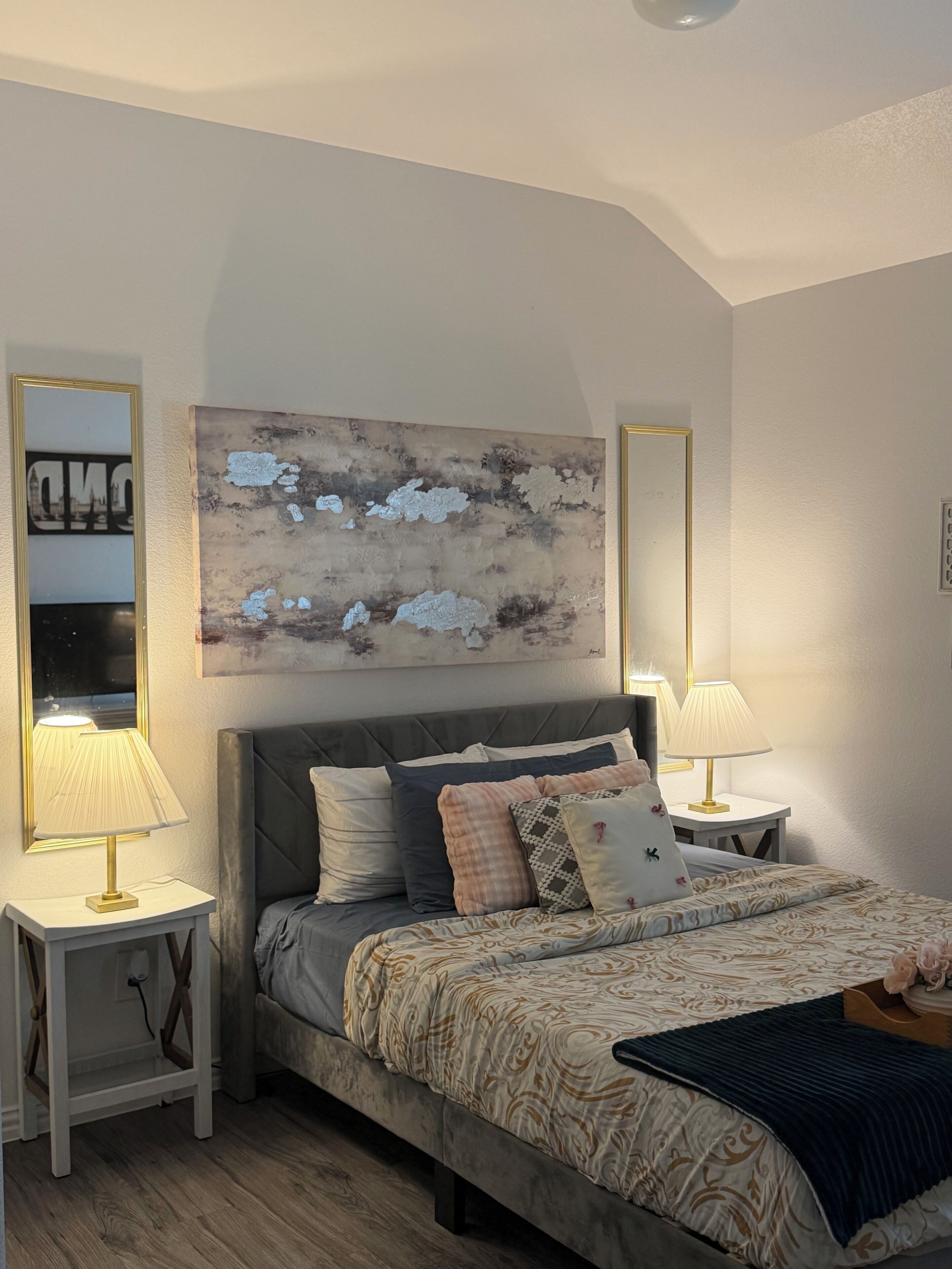 A bedroom with a bed, two bedside lamps, a large abstract painting, and a mirror on the wall.