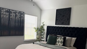 Une chambre à coucher avec une tête de lit capitonnée, un grand lit, un ventilateur de plafond, une fenêtre avec des stores et une œuvre d’art accrochée au mur.