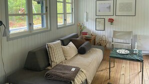 A cozy room with a sofa, a small table, and a chair. There are windows with curtains, a wall with framed pictures, and a wooden floor.