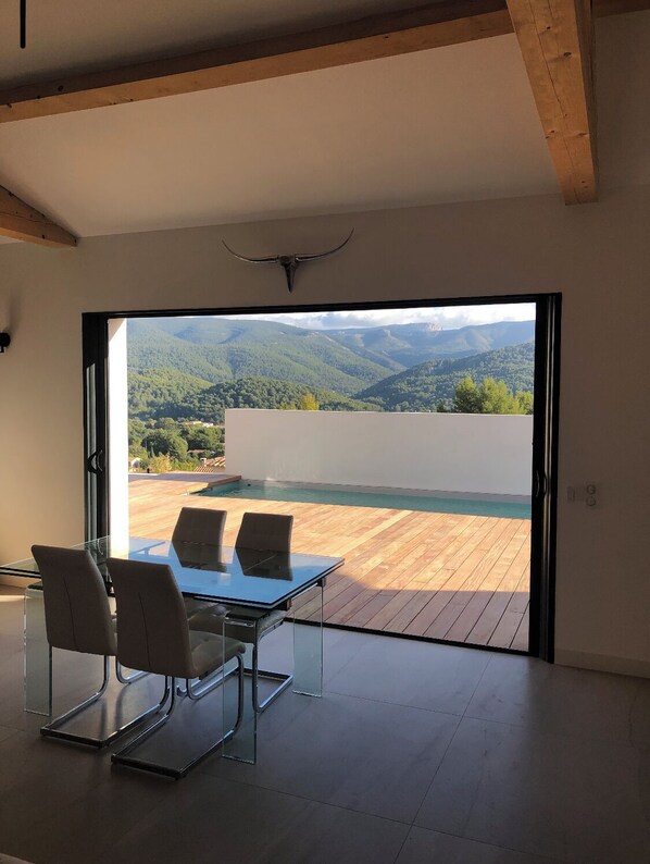 Ein modernes Zimmer mit Glastisch und -stühlen, einer Holzbalkendecke und einer großen Schiebeglasdoor zur Terrasse mit Pool und Blick auf eine Berglandschaft.