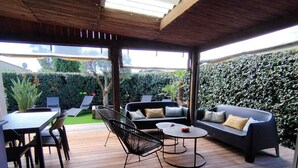 Een buitenterras met een houten dek, een bank, een eettafel met stoelen en een kleine ronde tafel.