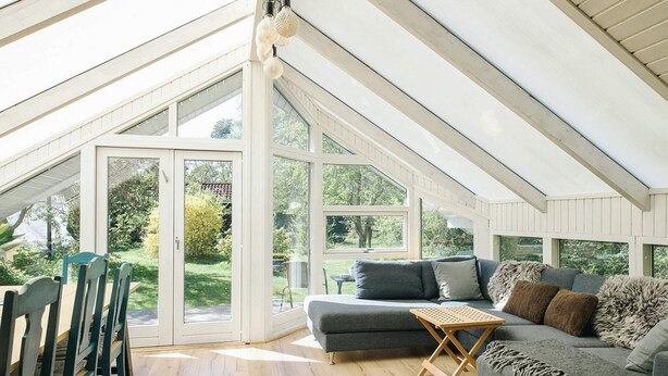 A conservatory with a high ceiling, a grey sofa with multiple cushions, a wooden table, and a dining area with chairs. - 4 Star Holiday Home in Gorlev (Gørlev)