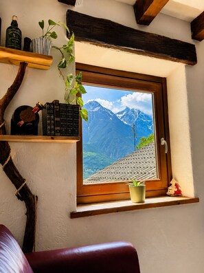 Una ventana con vistas a la montaña, un estante de madera con libros y botellas, una planta en maceta y un sillón de cuero rojo.