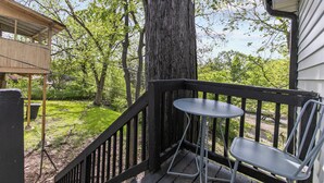 Eine Holzterrasse mit einem Metalltisch und Stuhl, umgeben von Bäumen und einer Rasenfläche.