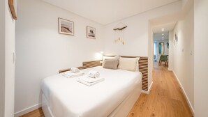 Quarto com cama grande, cabeceira de madeira, duas imagens penduradas na parede, um adorno suspenso e um corredor que dá para outro cômodo.